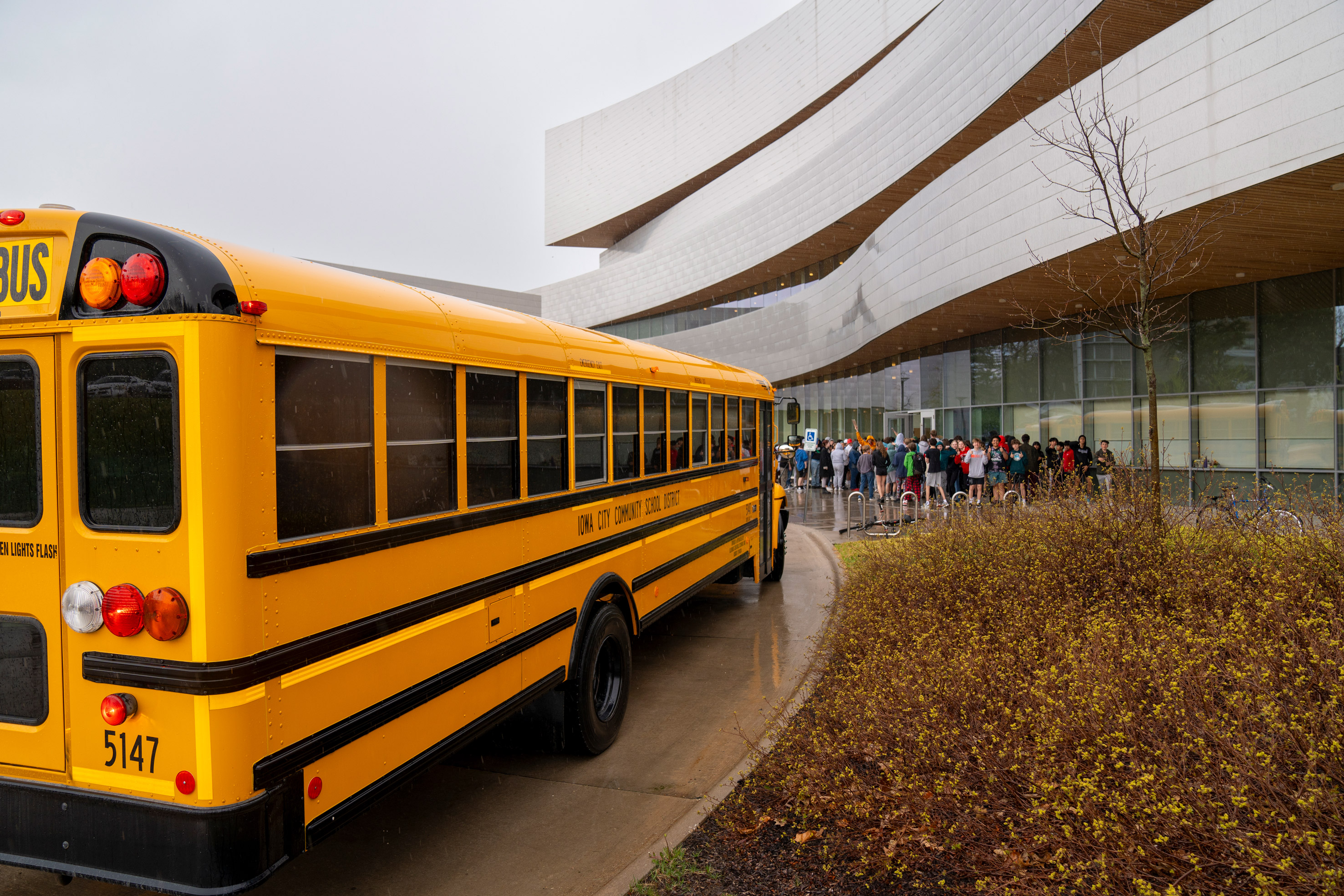 A yellow school bus and 8th grade students outside Hancher Auditorium on a gray, drizzly day