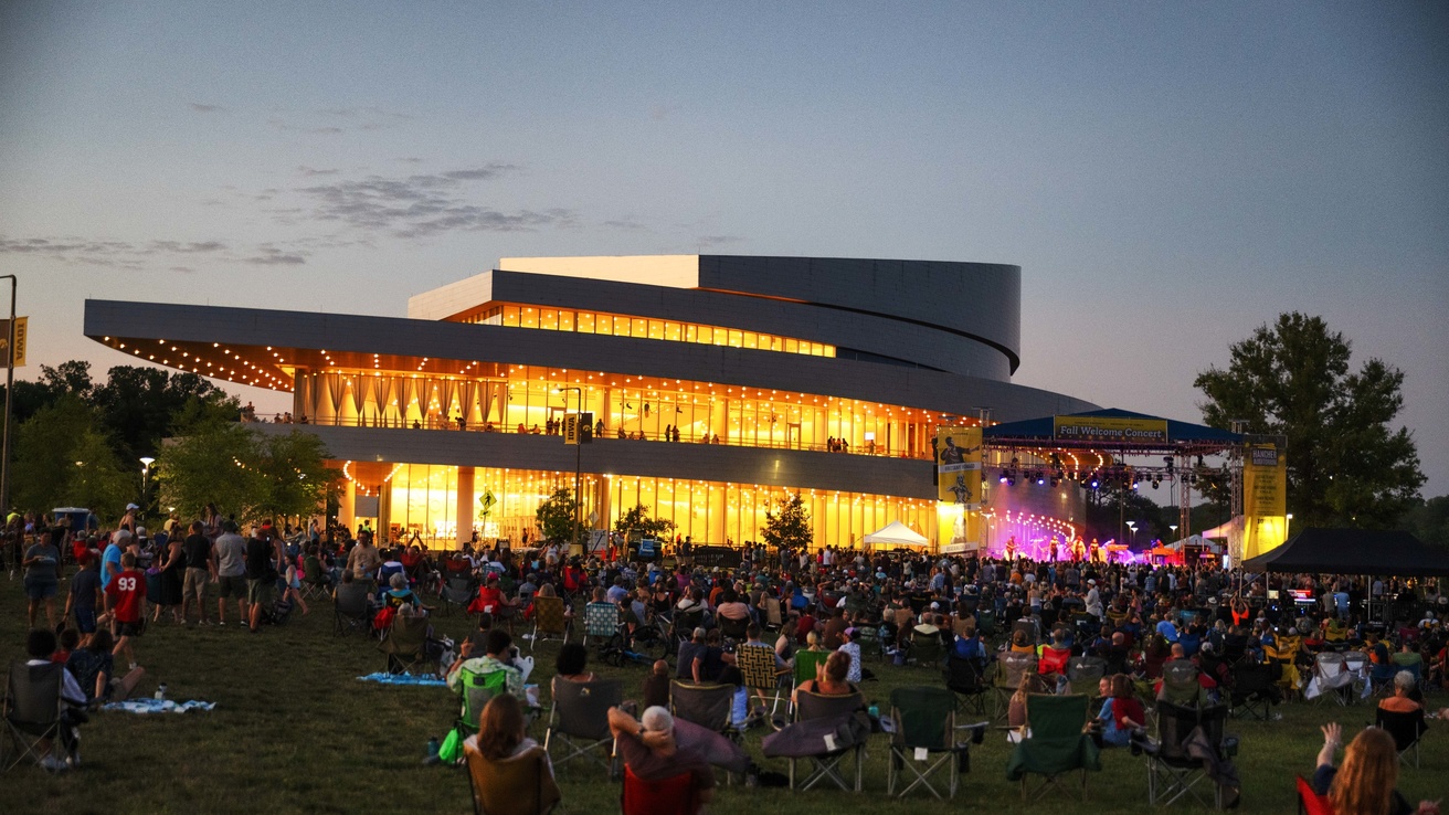 A crowd sits and stands outdoors on the Hancher Green, watching the Fall Welcome Concert on an outdoor stage set up in front of Hancher Auditorium.